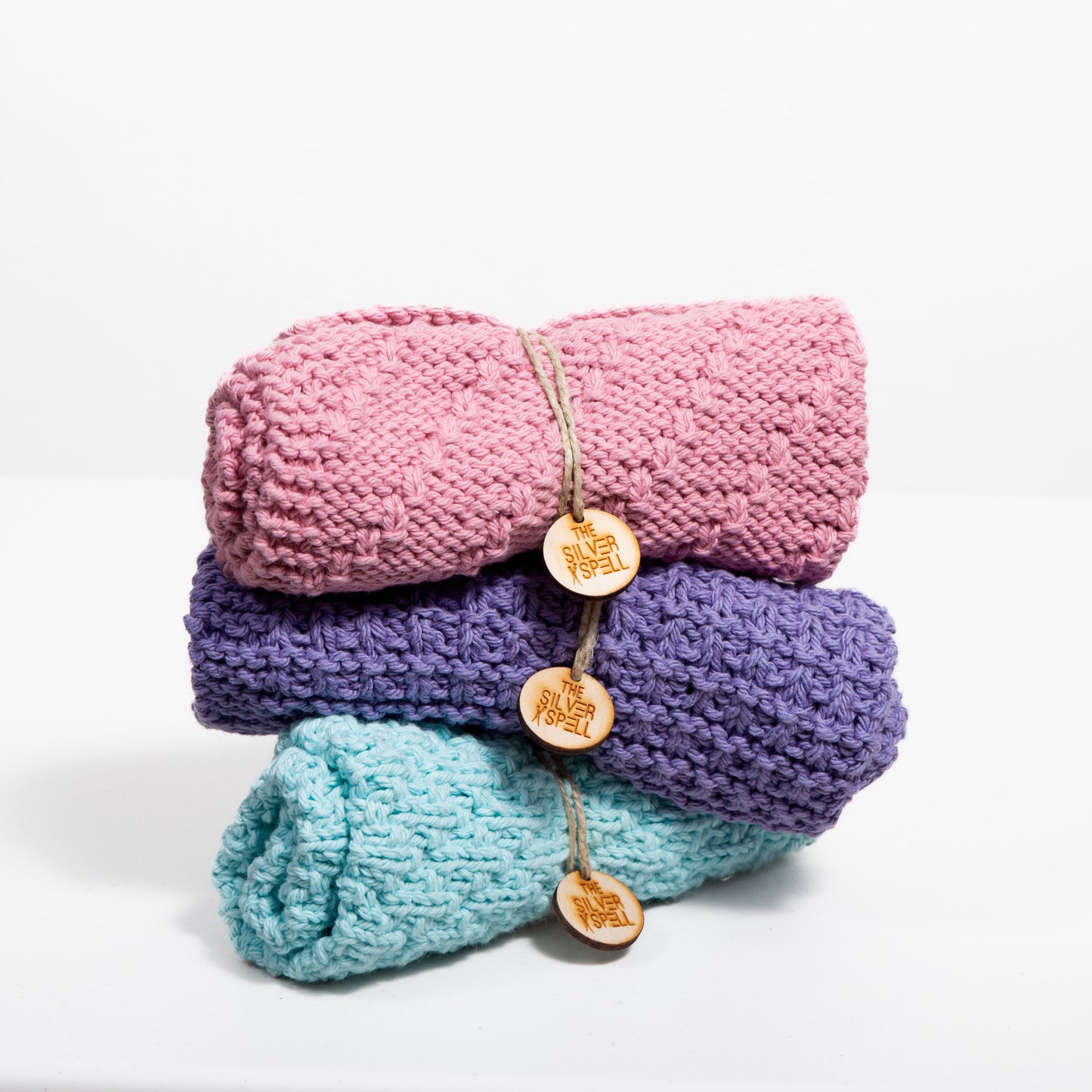 Three folded cotton towels in pink, purple, and blue, each with a brand tag attached.