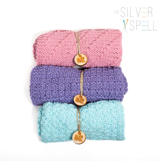 Three folded cotton towels in pink, purple, and blue, each with a brand tag attached.