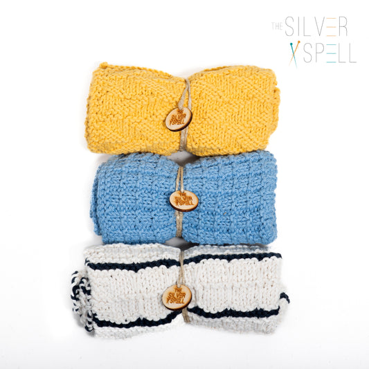 Three folded towels in yellow, blue, and white with black stripes, each featuring a button detail and branded tag on the blue towel.